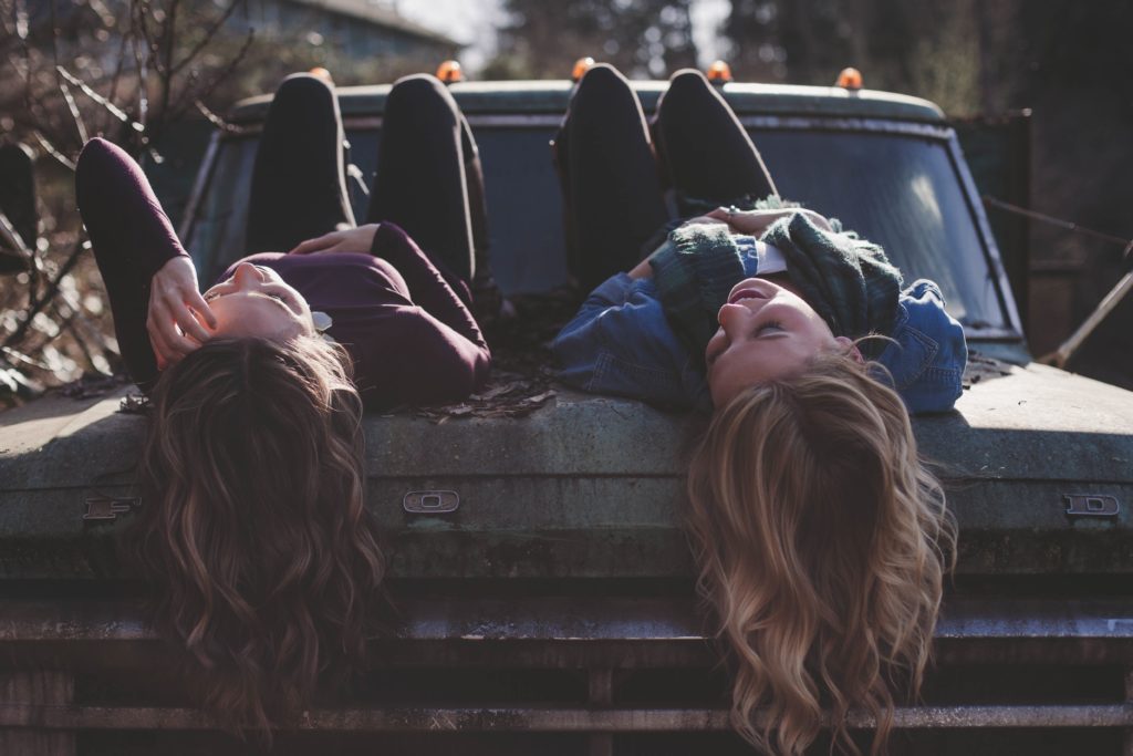 Two college age women sitting on a car hood, talking and looking at the sky.