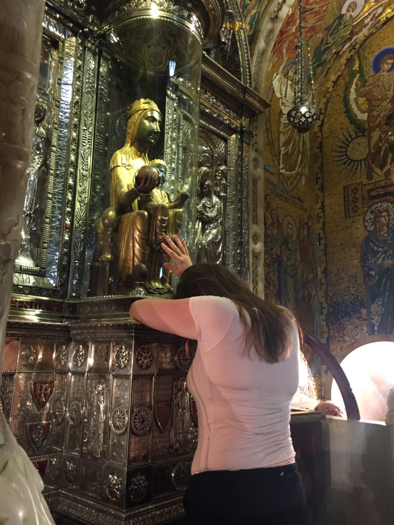 Woman rests hand on glass surrounding statue of black madonna.